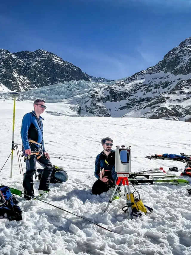 Zwei Forschende, einer stehend, einer kniend, schauen auf einen terrestrichen Laserscanner, der Teil eines Messsettings in einem verschneiten Alpengelände ist.